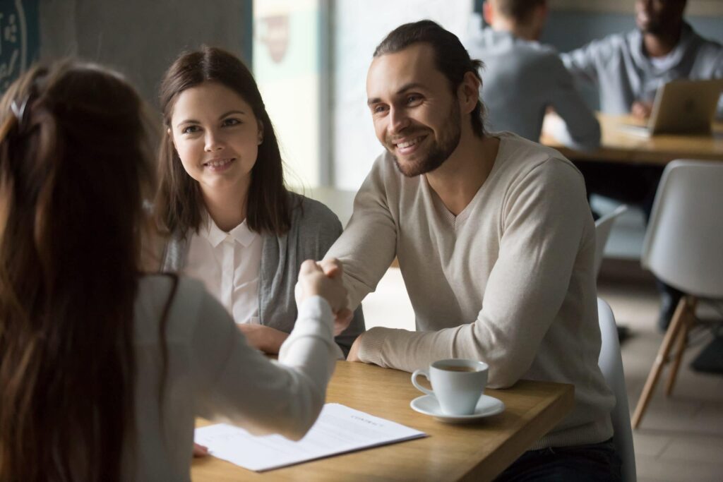 Three people sit at a table in a cafe; a woman shakes hands with a man across the table while another woman observes. A document and coffee cup are on the table.