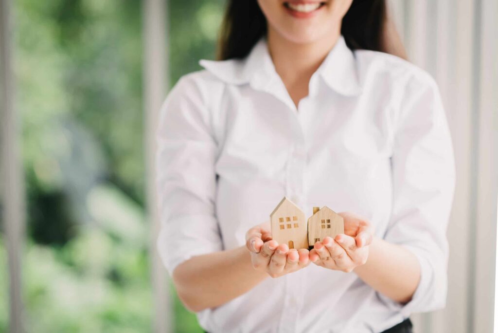 Woman in a white shirt holding two small wooden house models in her hands, smiling, with a blurred green background.