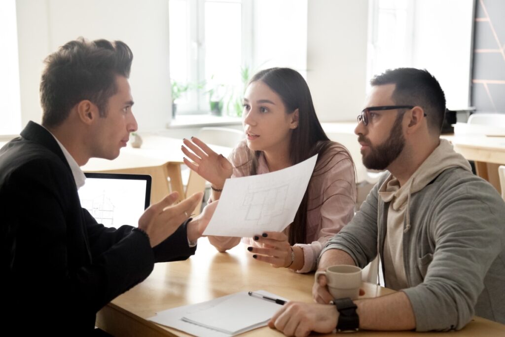 Three people sit at a table having a discussion, with one woman holding documents and gesturing while the other two listen. A laptop and coffee cup are on the table.