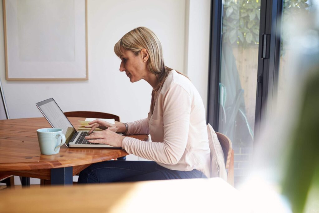 Woman sitting at a wooden table using a laptop, with a blue mug and notepad beside her, in a bright room with large windows.