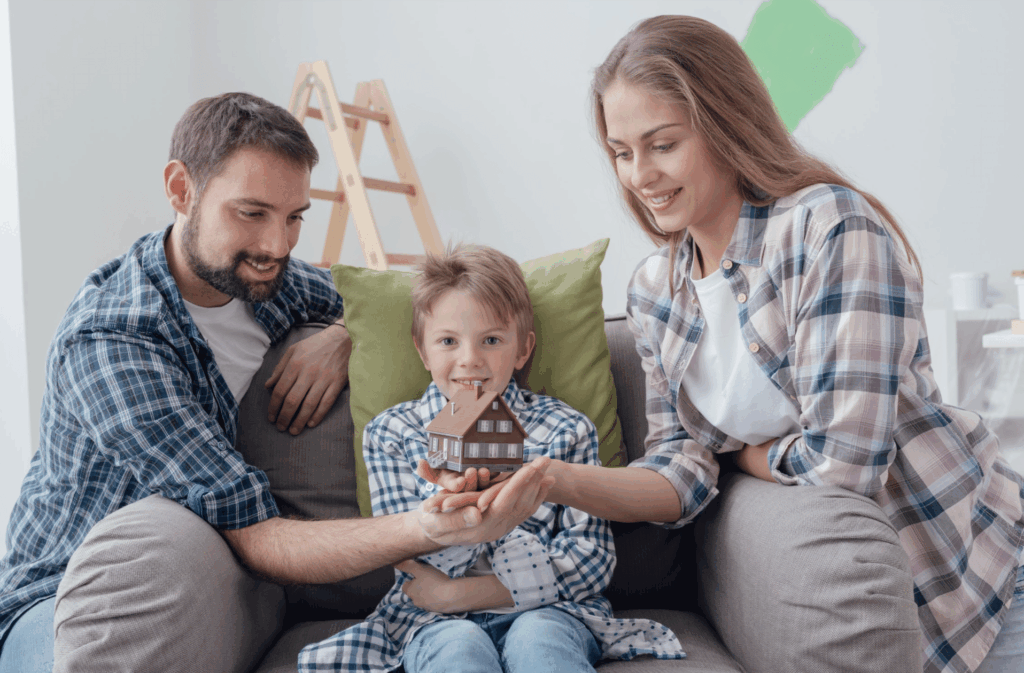 A man, a woman, and a child sitting on a couch hold a small model house together, with a ladder and green paint on the wall in the background.