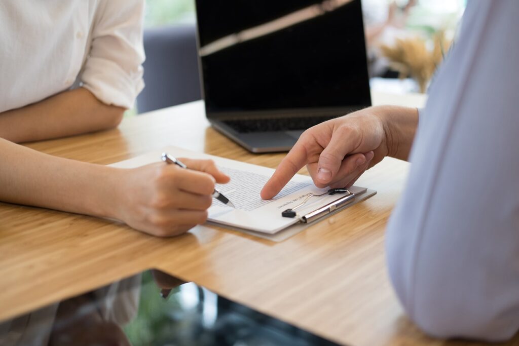 Two people sit at a desk with a laptop, reviewing and discussing a document on a clipboard; one person points while the other holds a pen.