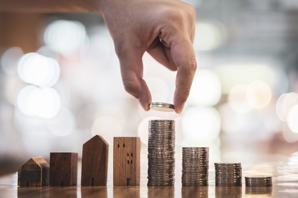 A hand places a coin on top of a stack of coins arranged in ascending order, with small wooden house models beside the stacks on a reflective surface.
