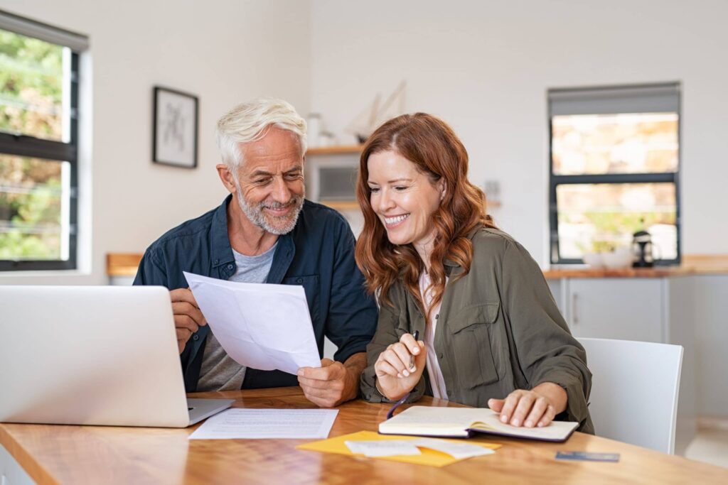 Smiling man and woman sit at a table with a laptop, paperwork, and notebooks, appearing to review documents together in a bright home office.