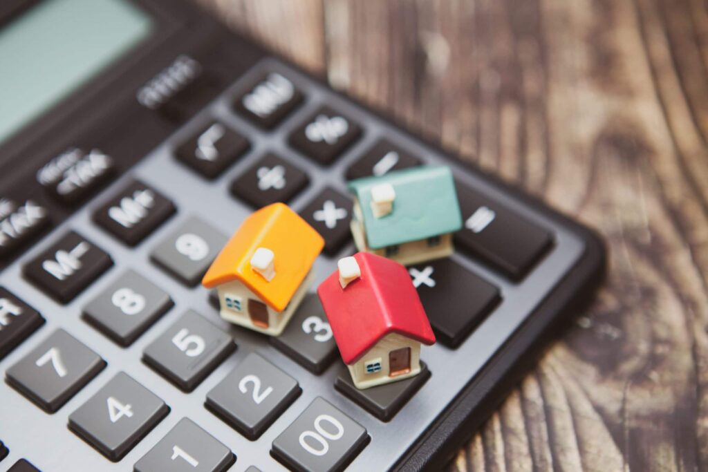 Three small model houses are placed on the buttons of a black calculator, all set on a wooden surface.