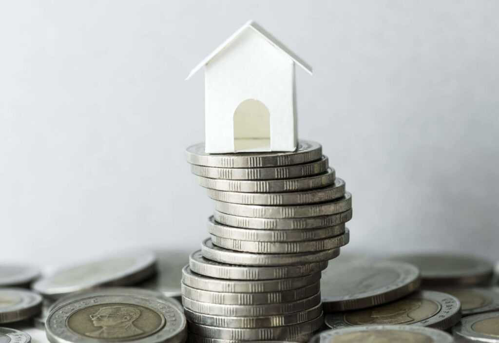 A small white house model sits on top of a stack of coins, symbolizing real estate investment or property value.