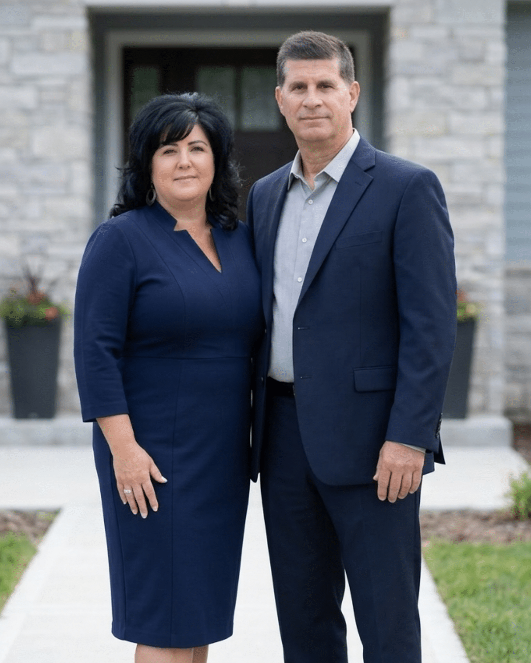 A woman and a man stand side by side in front of a stone building, both dressed formally in navy blue outfits, looking at the camera.