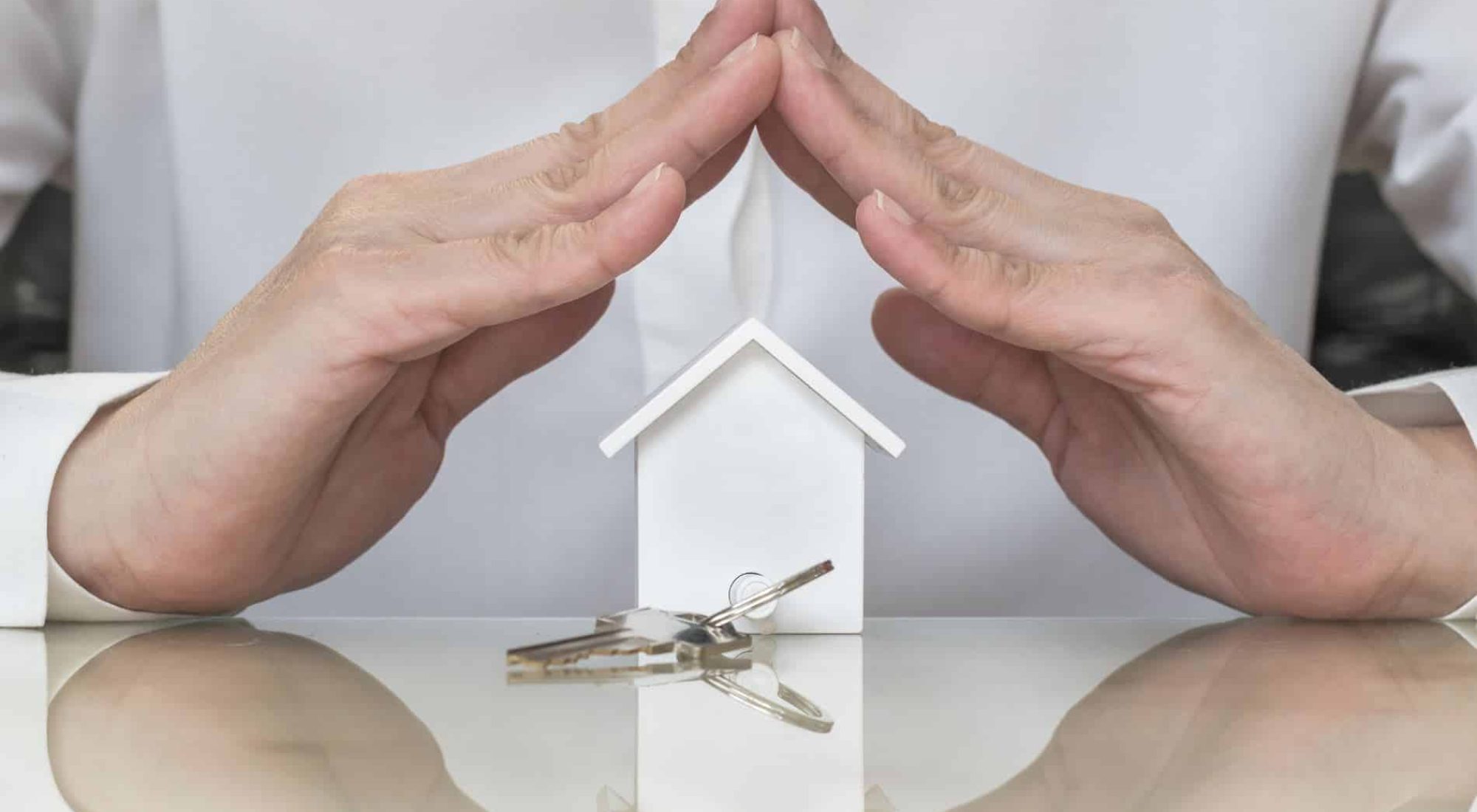 A person’s hands forming a roof shape over a small house model, with a set of keys placed on the reflective surface in front.