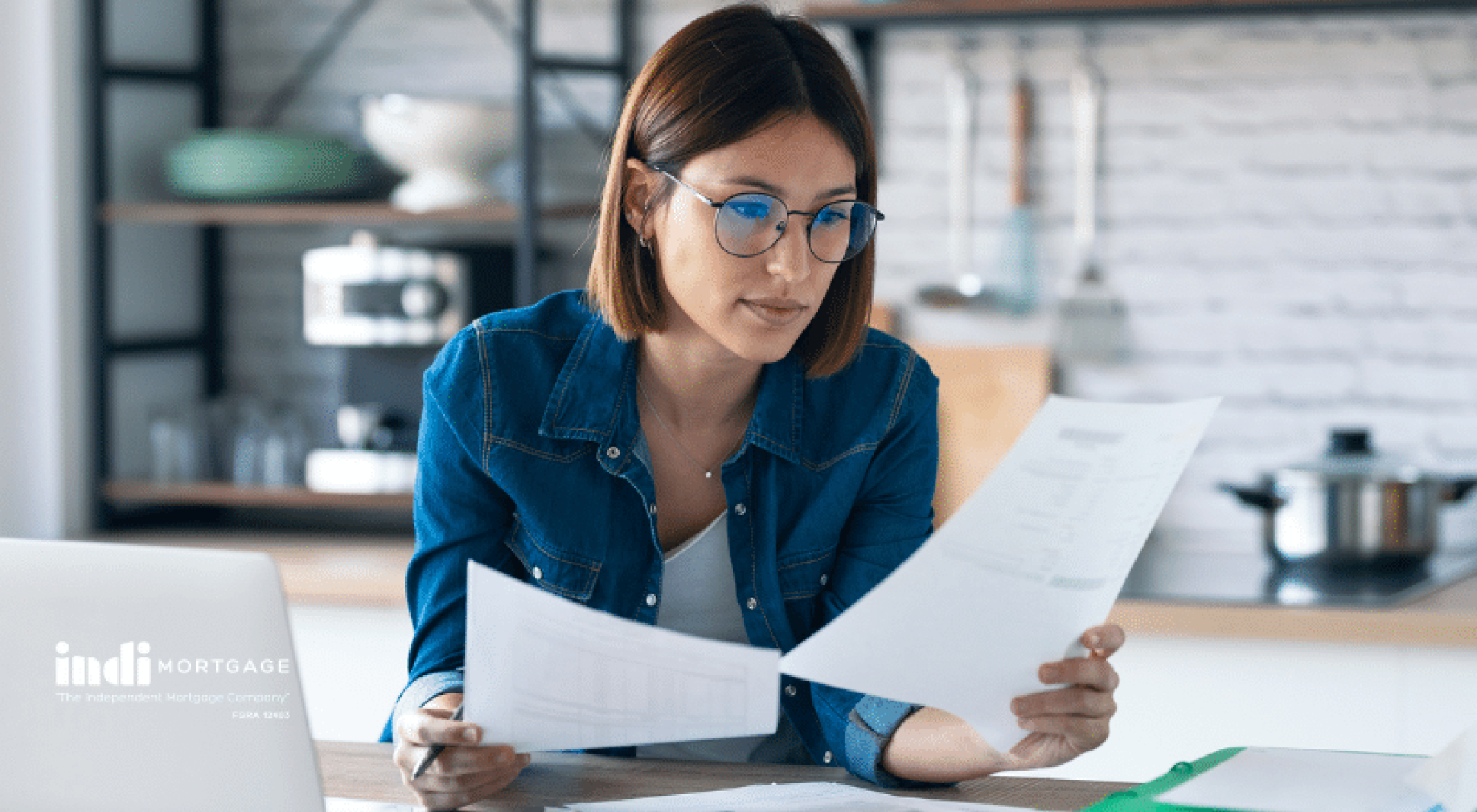 Woman wearing glasses reviews documents at a kitchen table, with a laptop and clipboard in front of her.