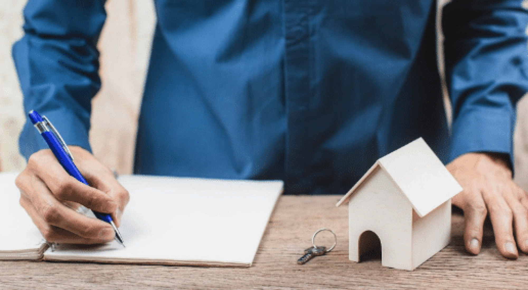 Person writing on a notebook at a desk with a small house model and keys placed nearby.