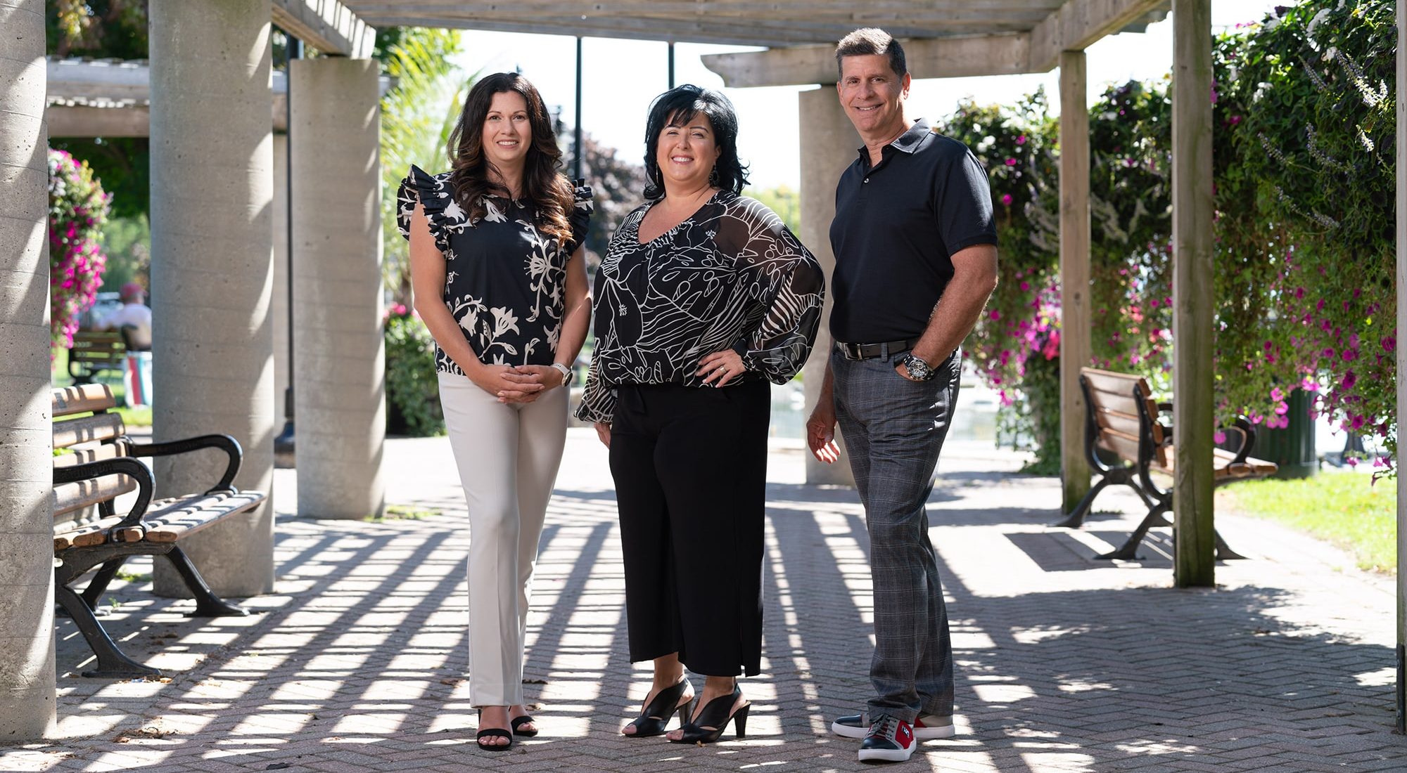 Three adults stand side by side outdoors under a pergola on a sunny day, with benches and hanging flower baskets in the background.