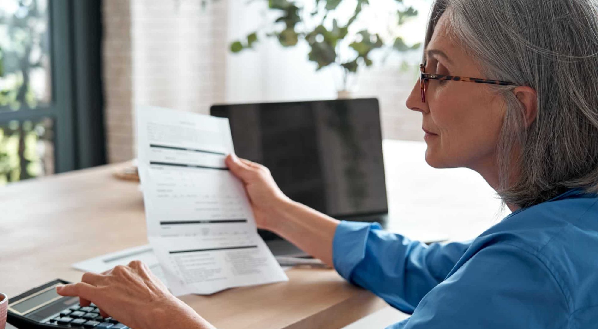 Older woman with gray hair and glasses reviews a document while using a calculator at a desk with a laptop and coffee cup.