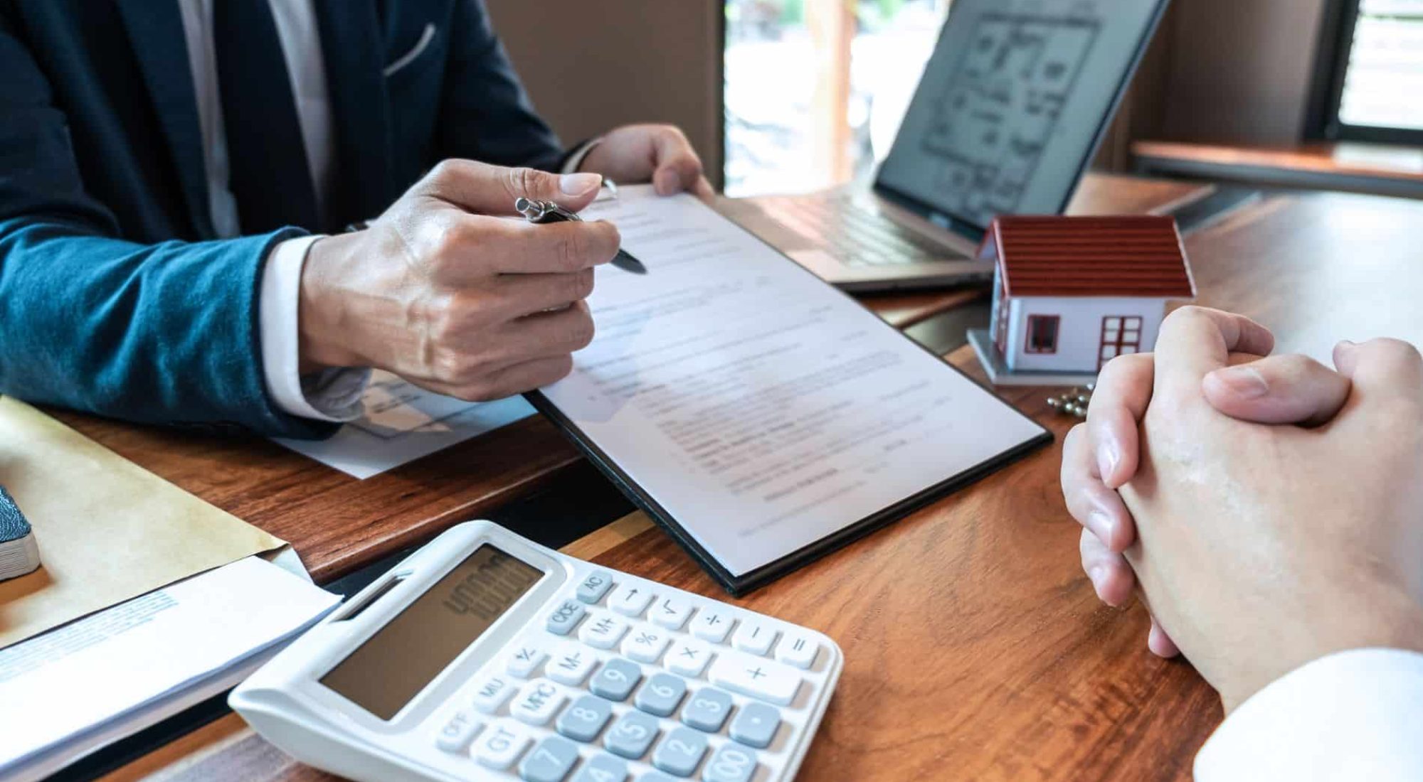 Two people sit at a desk with documents, a calculator, a laptop, and a small model house, discussing paperwork related to real estate or financial transactions.